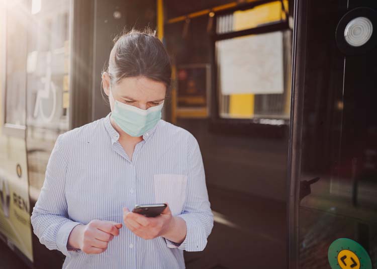 Frau mit einer Gesichtsmaske steht vor Bus und hält Mobitelefon © Nils - stock.adobe.com Frau mit einer Gesichtsmaske steht vor Bus und hält Mobitelefon